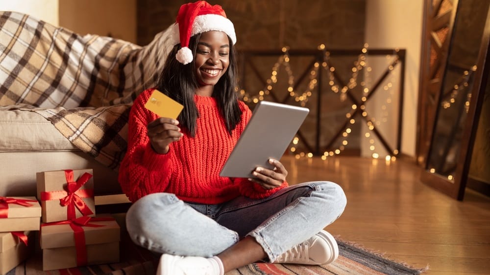Happy black lady in Santa hat using digital tablet and credit card to buy Christmas gifts on web from home