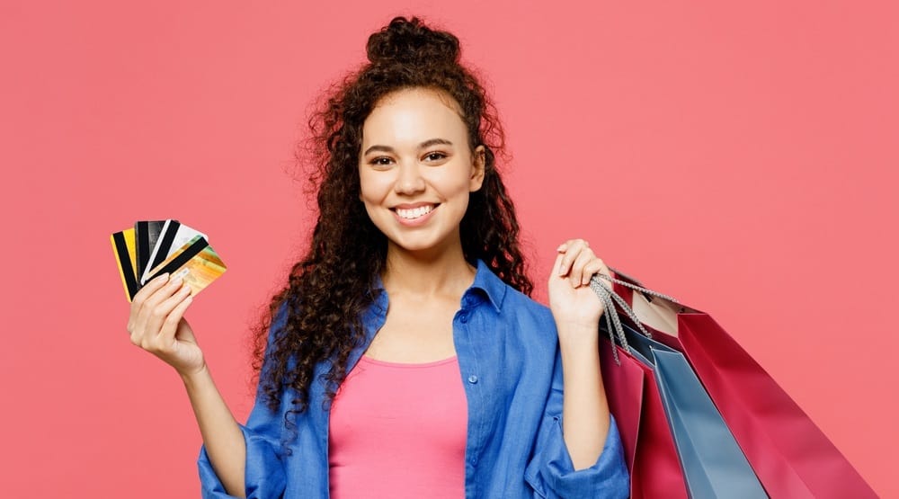 Wanita muda yang tersenyum mengenakan pakaian kasual kemeja biru memegang tas paket kertas kartu kredit di tangan setelah berbelanja terisolasi di studio latar belakang merah muda pastel polos